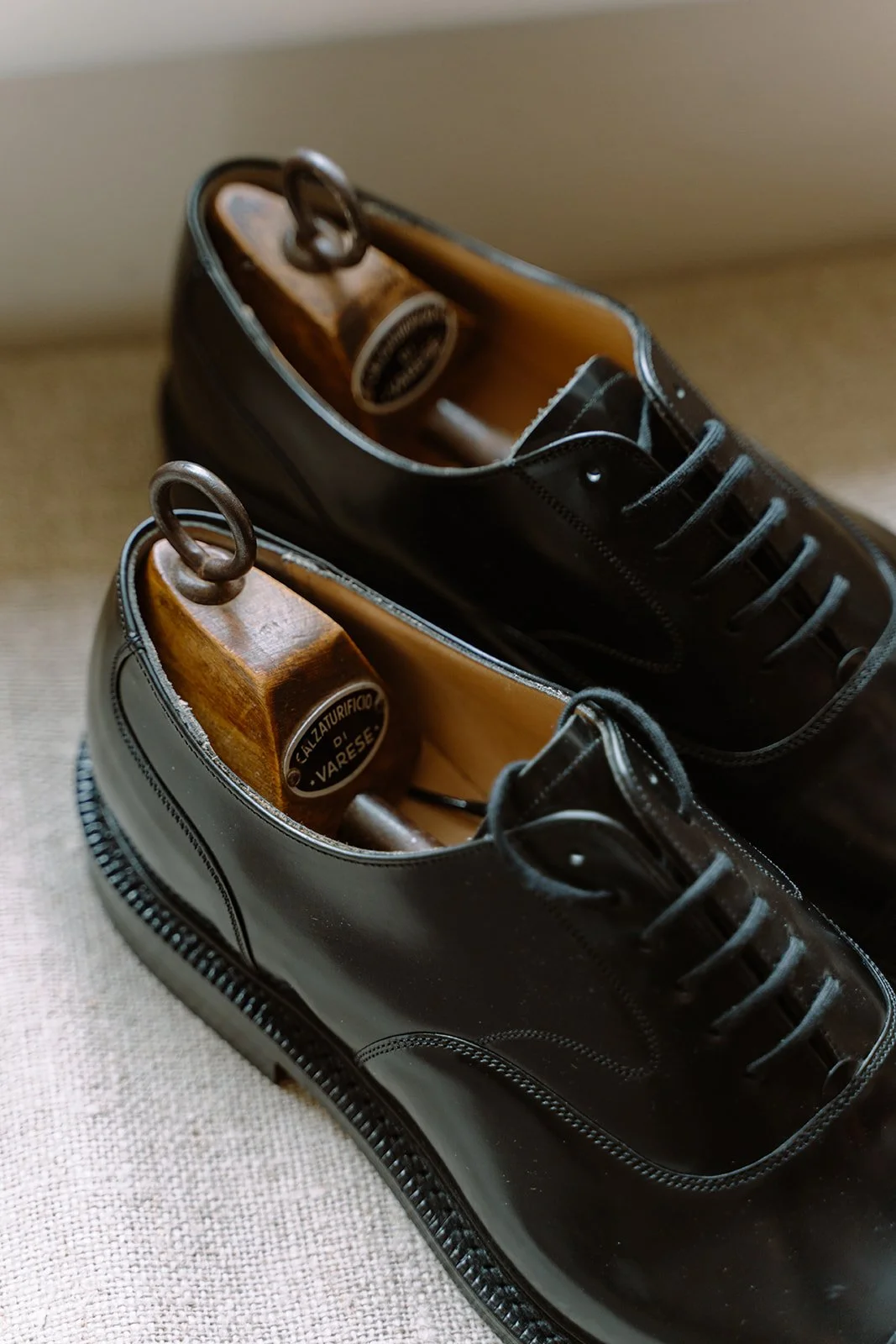 Pair of black leather Oxford shoes with wooden shoehorns inside, placed on a beige surface.