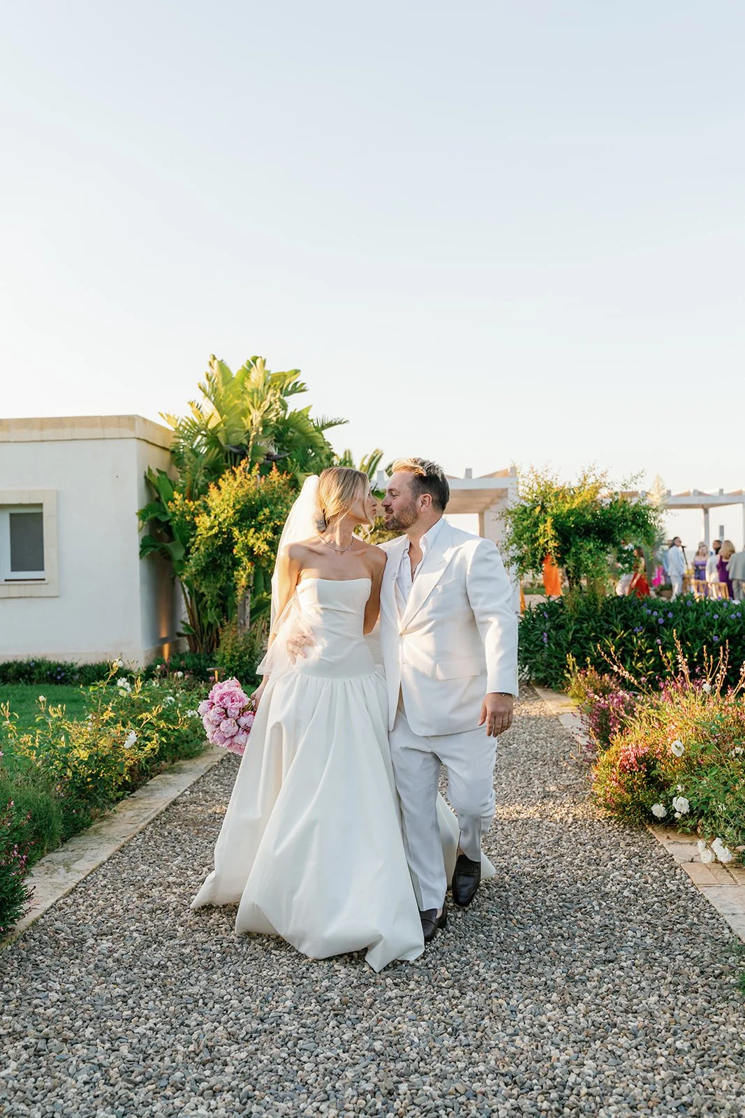 A bride and groom walking together outdoors, surrounded by greenery and flowers, during their wedding celebration.