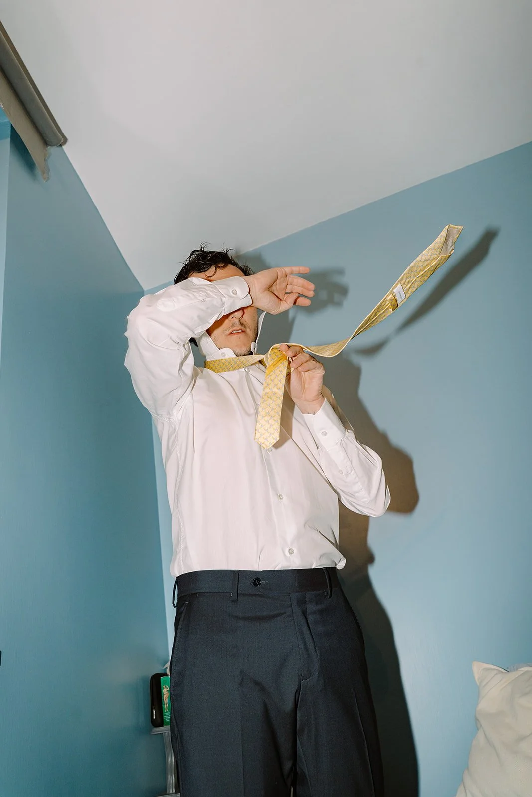 A man in a white shirt and dark pants is throwing a necktie into the air while covering his eyes with his arm in a room with blue walls.