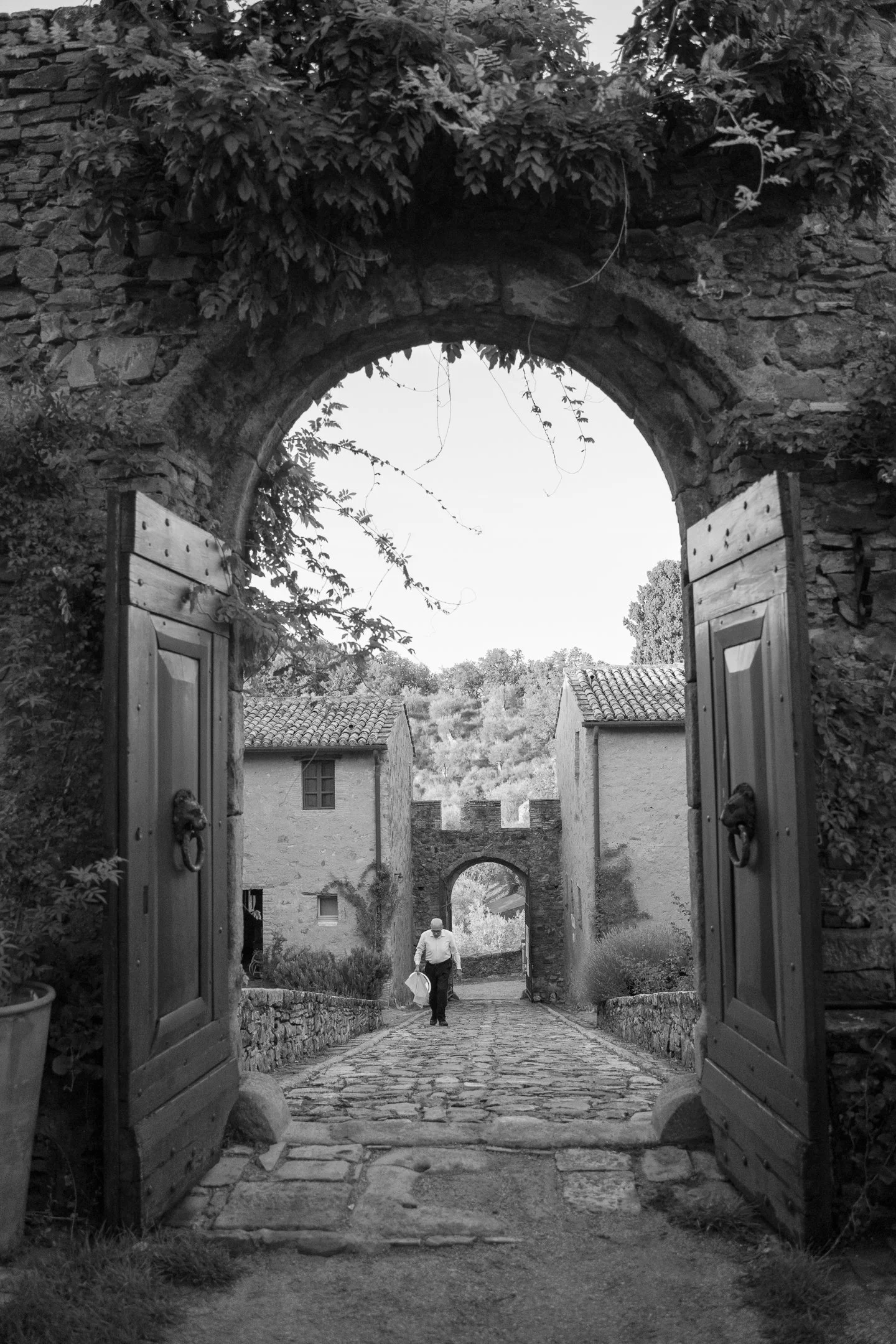 A black-and-white photograph of an open wooden gate leading to a stone-paved pathway lined with stone walls and historic buildings, with a man walking down the pathway carrying a bag, and a stone archway in the background.