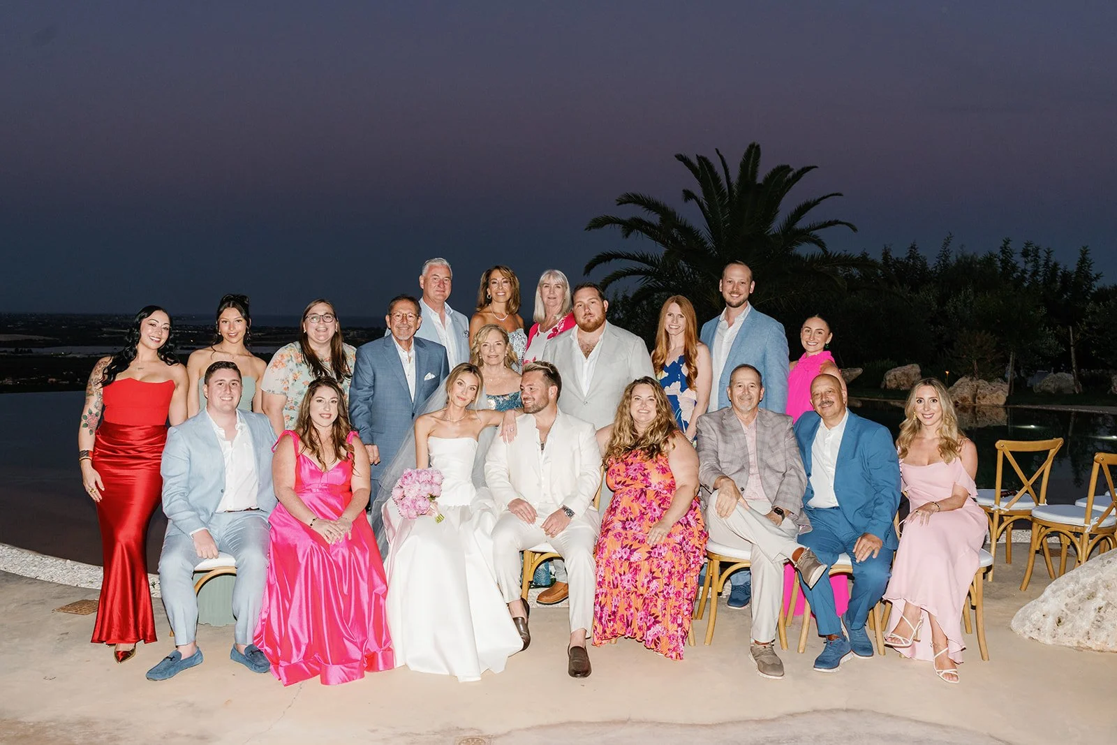 Group of people dressed in formal attire at a wedding or celebration outdoors during dusk, with a pool, stone deck, and a large palm tree in the background.