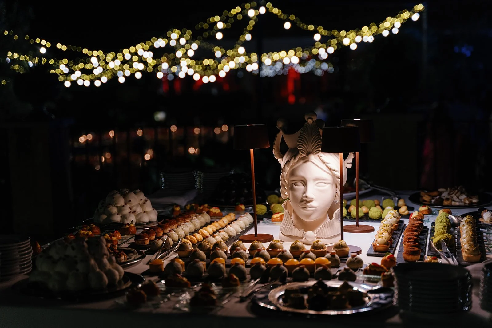 Dessert table featuring a large decorative head sculpture with lamps, surrounded by various pastries and sweets, illuminated by string lights in an outdoor setting at night.
