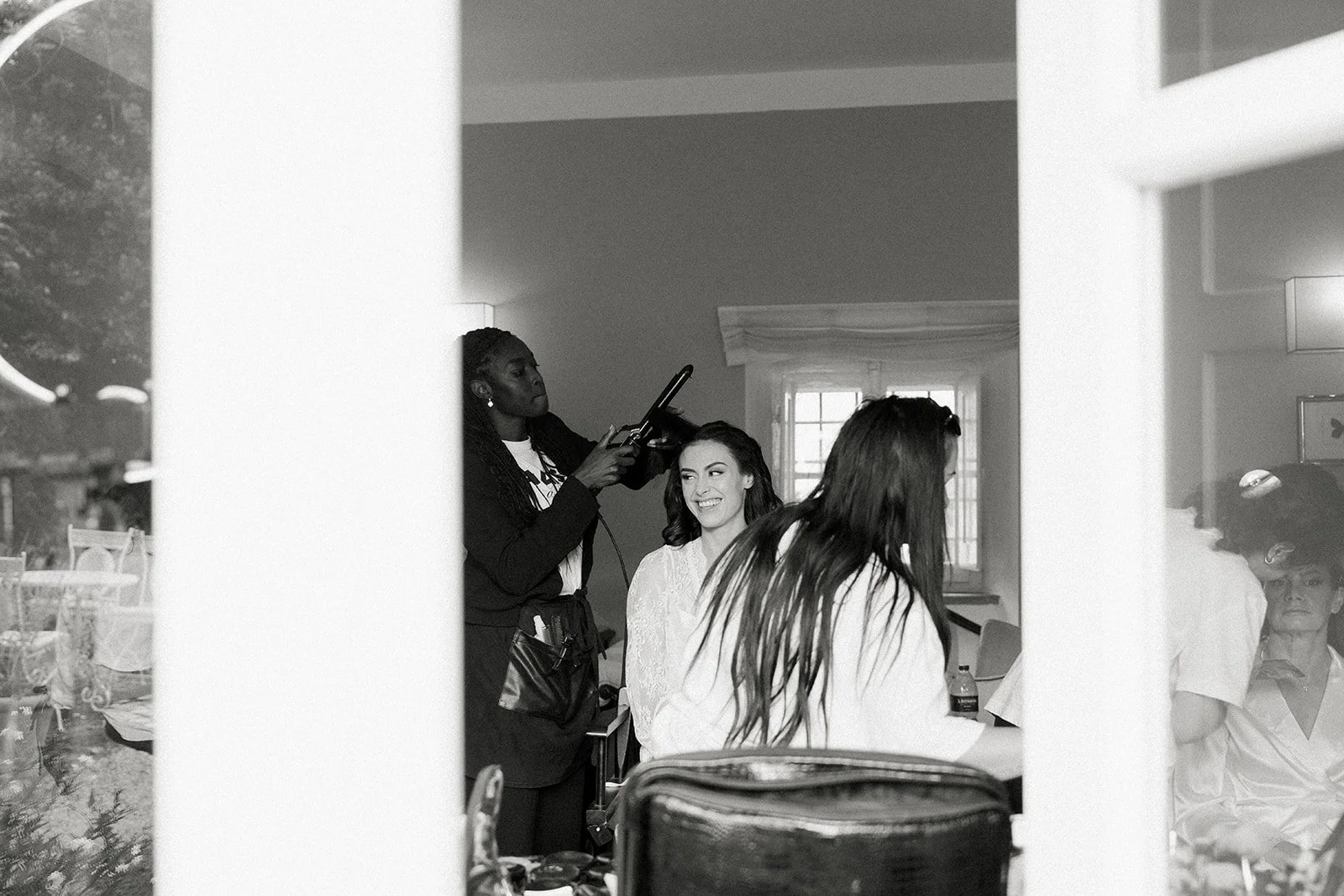 A woman getting her hair styled at a salon, smiling, with a hairstylist working on her hair, seen through a window.