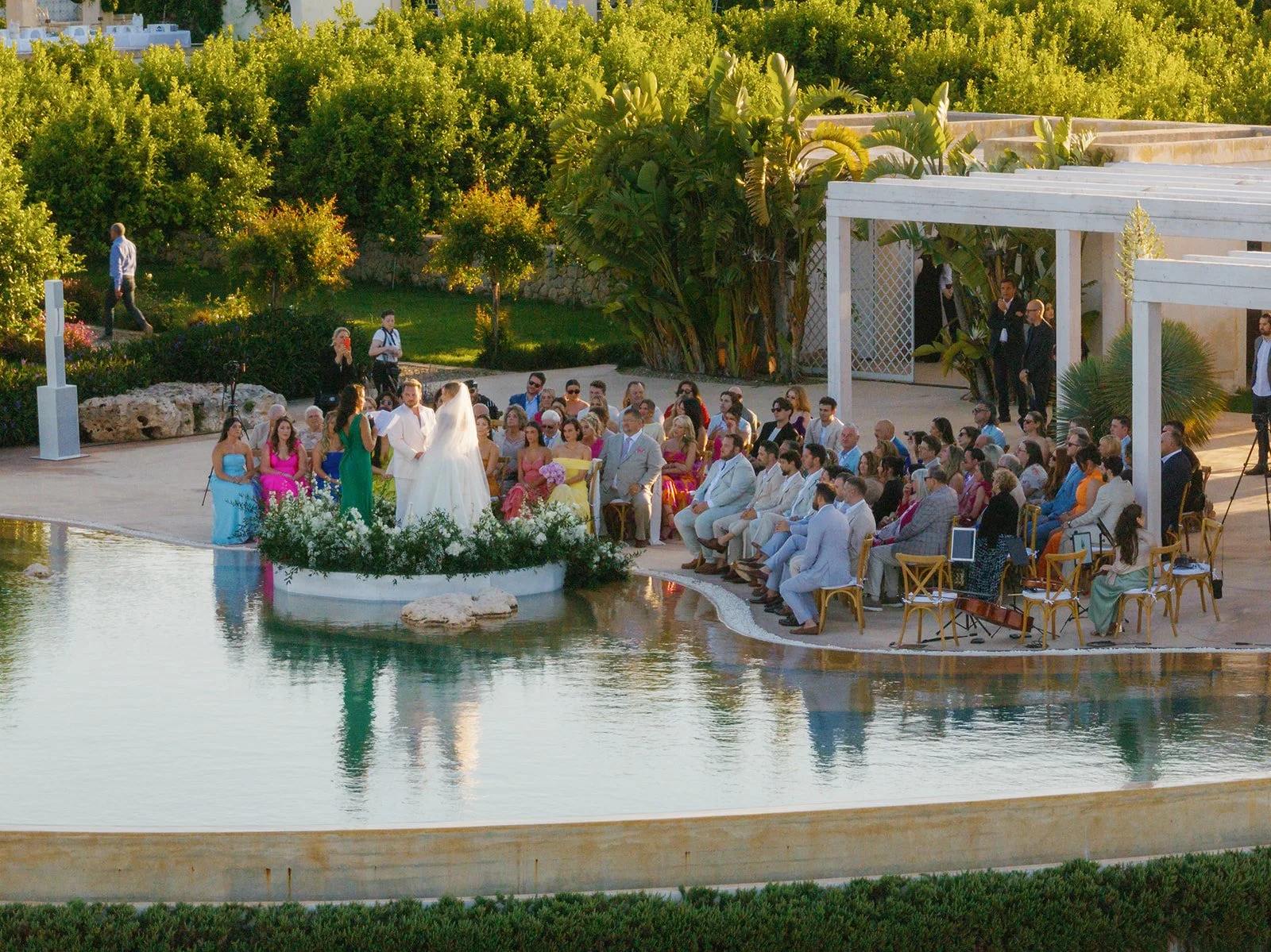 A wedding ceremony taking place outdoors beside a reflecting pool with lush greenery and trees in the background. The bride in a white gown and veil faces the officiant, while guests seated on chairs and some standing observe. Sunlight illuminates th