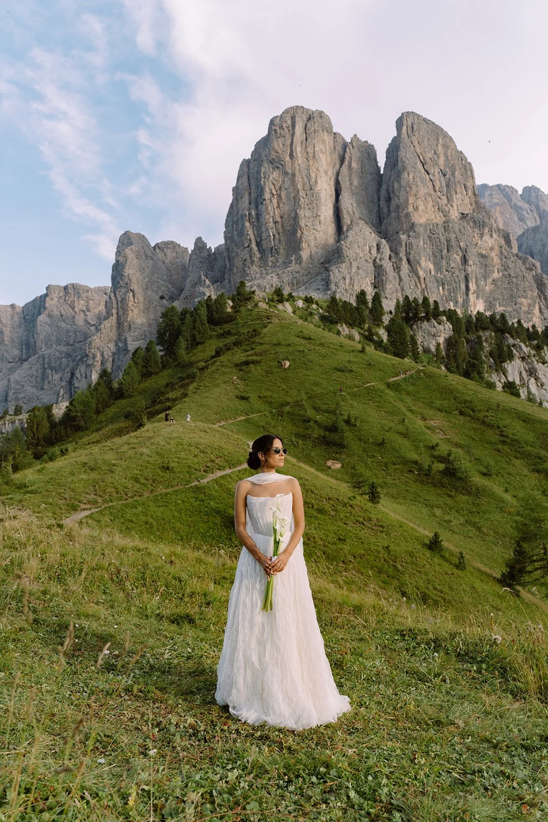 Woman in a white dress holding flowers standing on a grassy hillside with mountains in the background.