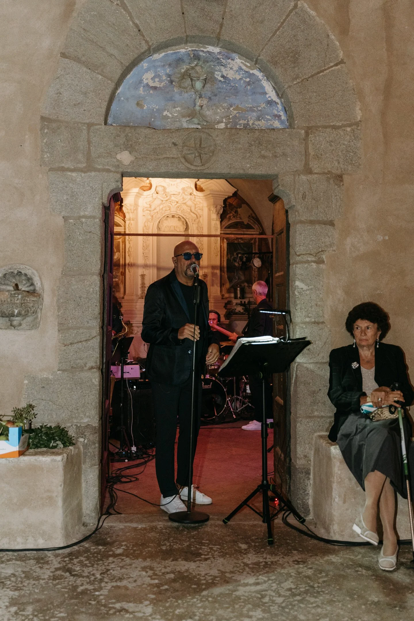 A man singing into a microphone during a live musical performance, with a band playing in the background, in a venue with rustic stone walls and an ornate interior visible through an arched doorway.