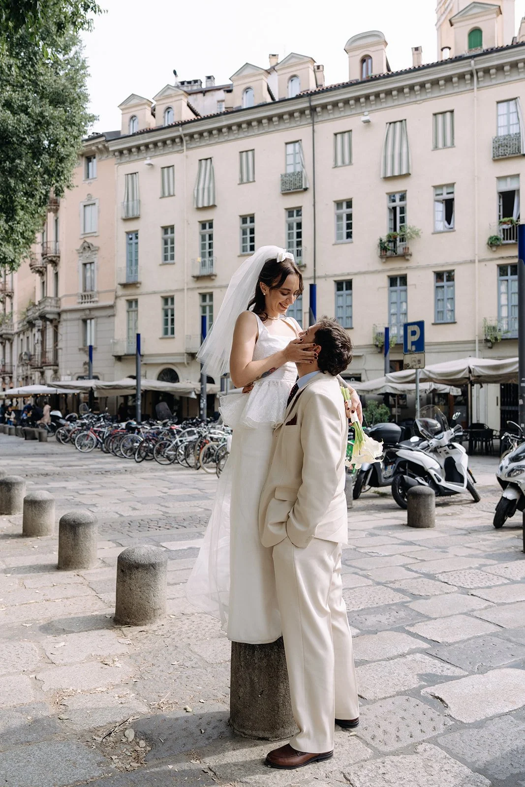 A couple dressed in wedding attire in an urban setting, with the woman standing on a stone post and the man holding her, smiling at each other, surrounded by parked bikes and scooters, with historic European-style buildings in the background.