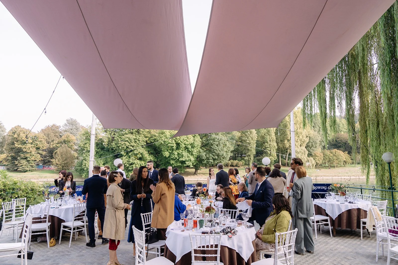 Outdoor gathering with people in formal attire, seated at decorated tables, under large pink canopy, near a body of water and lush green trees.