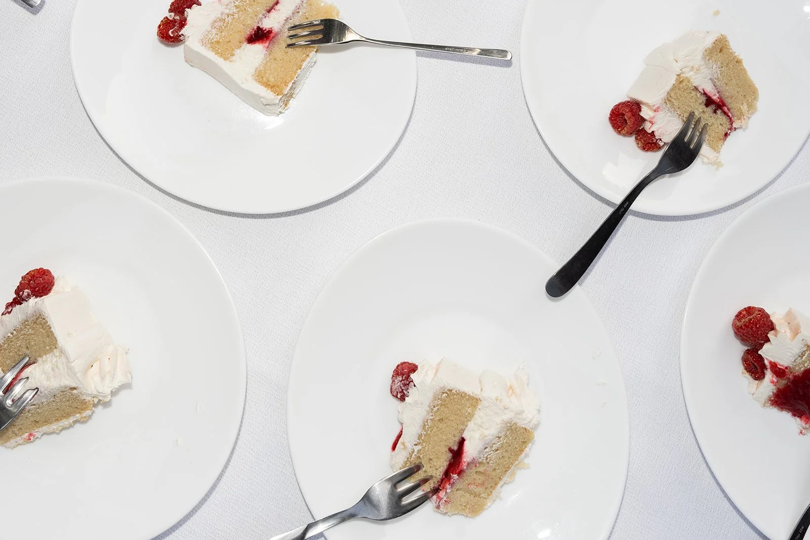 Four white plates with slices of cake topped with cream and raspberries, and forks resting on some of the plates, seen from above.