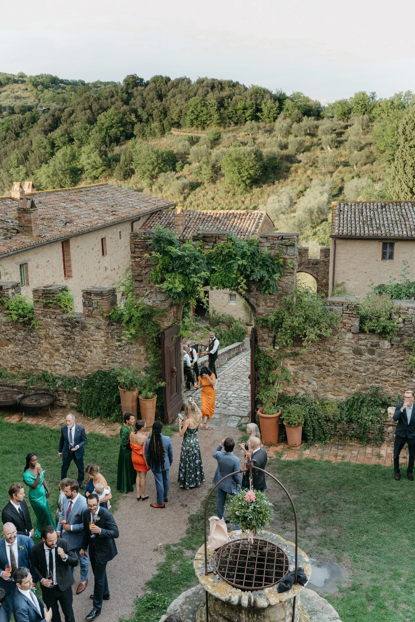 People gathered at a rustic outdoor wedding or party in a historic stone courtyard with lush greenery, potted plants, and scenic hillside in the background.