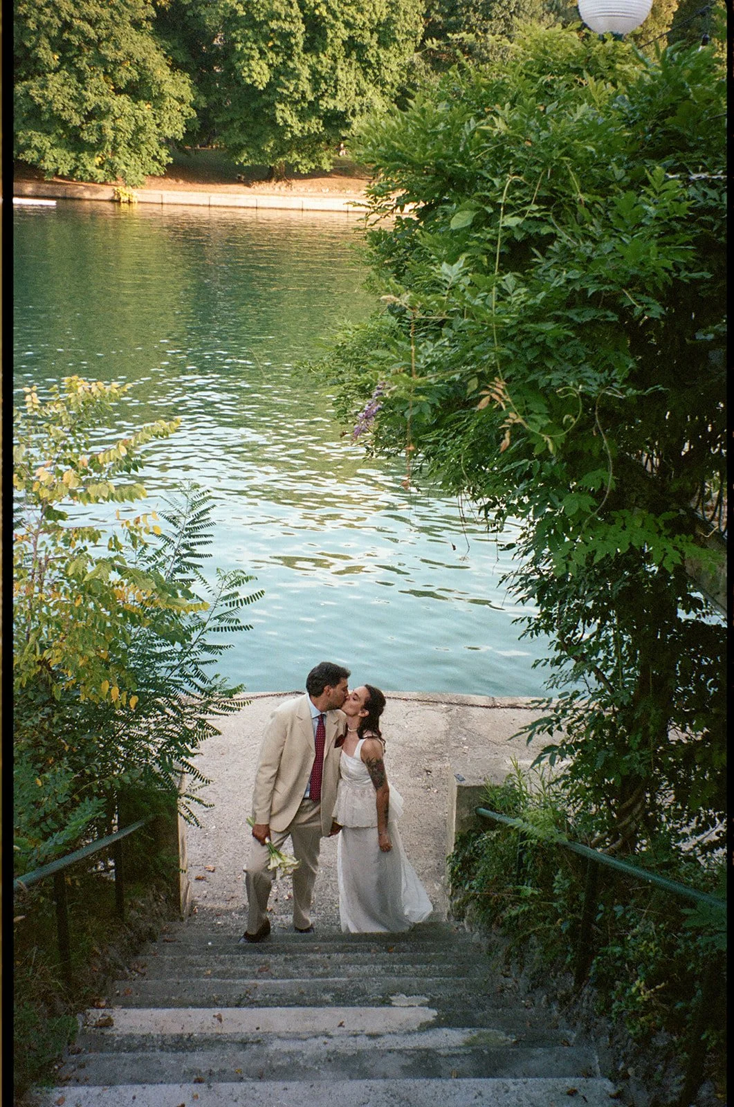 A newlywed couple kissing at the bottom of stone steps leading to a lake, surrounded by green foliage.