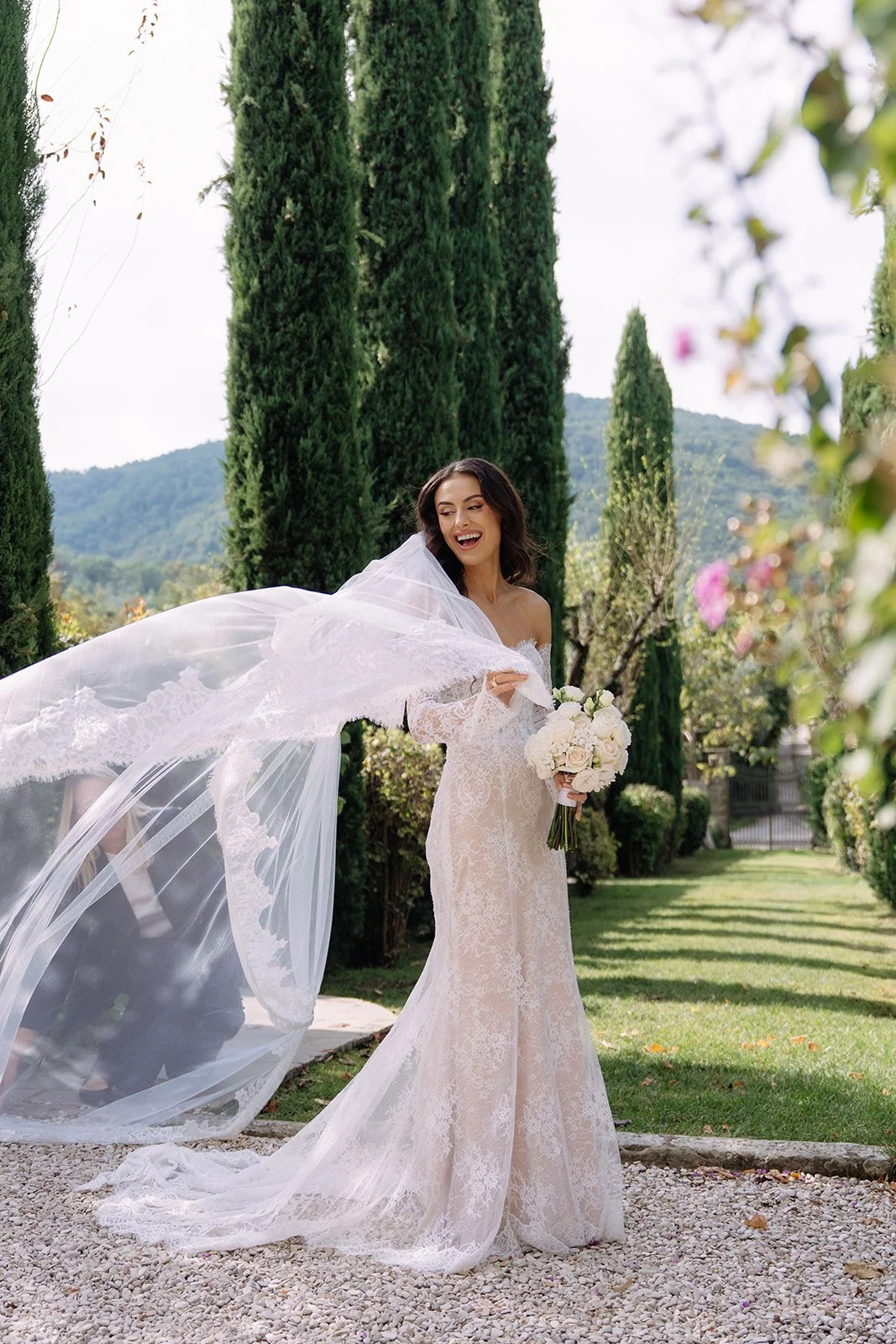 A bride in a lace wedding gown holding a bouquet of white roses and smiling outdoors among tall green cypress trees and mountains in the background.