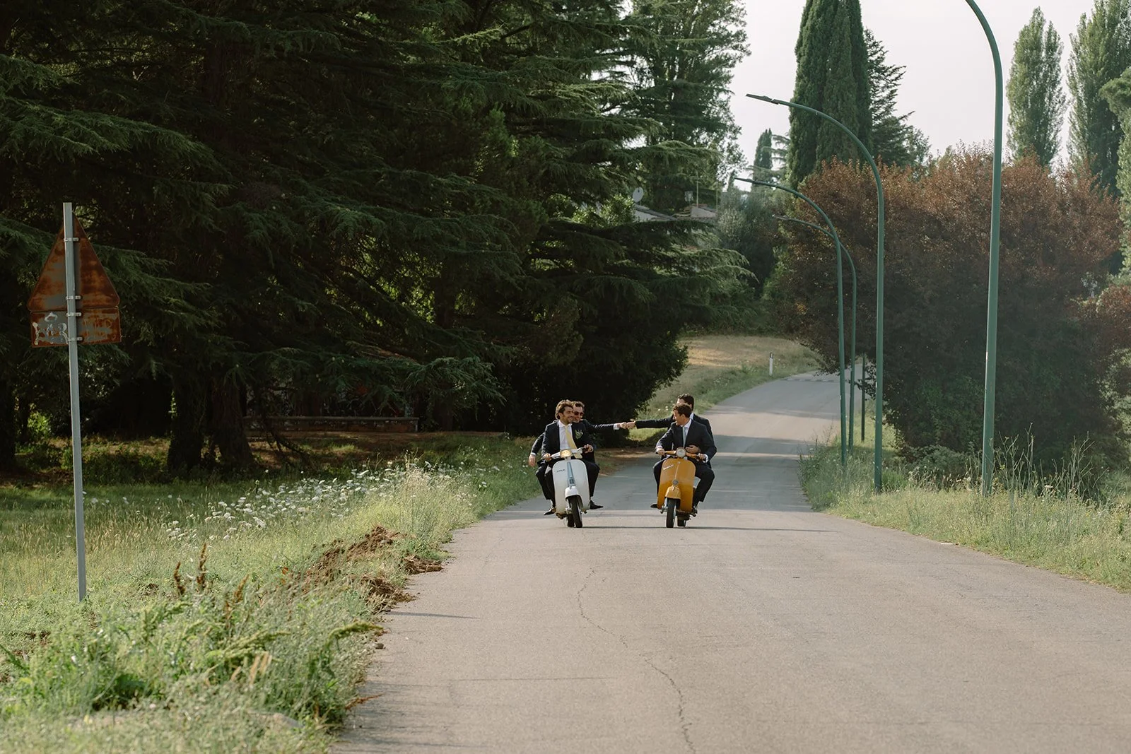 Three men in suits riding scooters on a rural road, with two of them reaching out and holding hands.