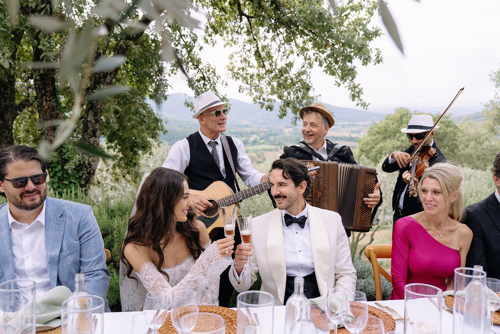 Group of people celebrating at an outdoor event, with musicians playing guitar, accordion, and violin in the background.
