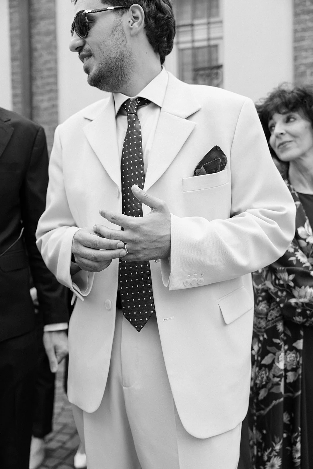 A man in a white suit and polka dot tie, wearing sunglasses, standing outdoors, smiling, with a woman in a floral dress in the background.