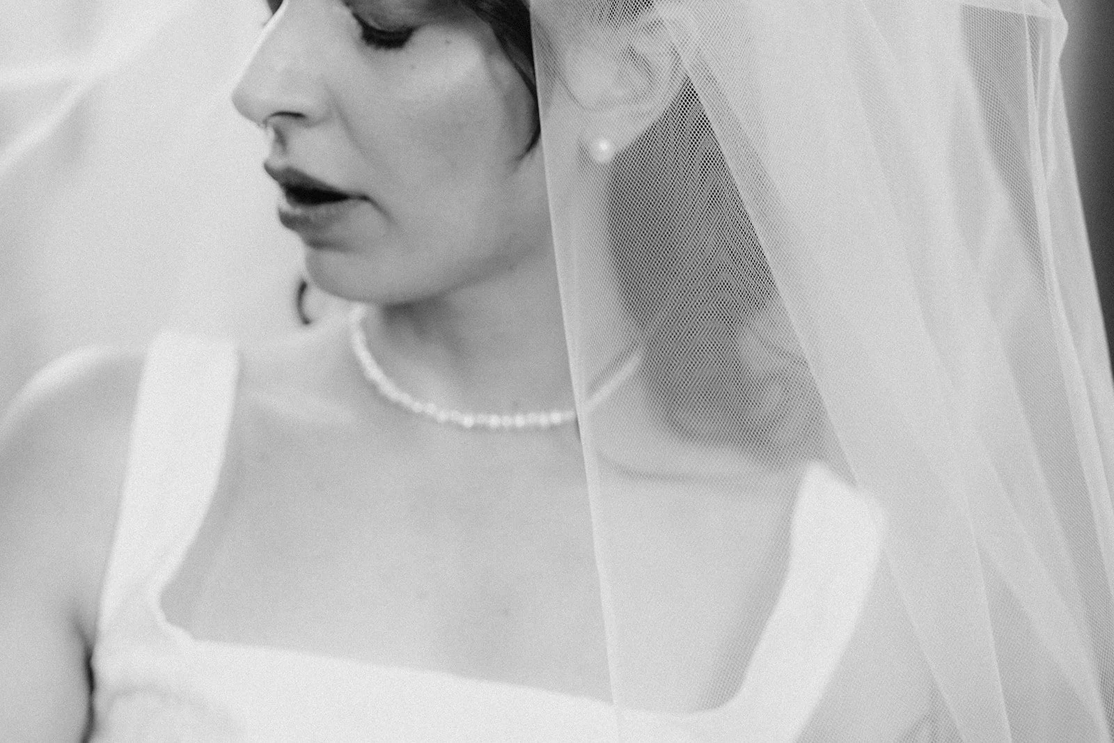 Black and white close-up of a bride looking down, wearing a wedding dress, pearl necklace, and a veil over her face.