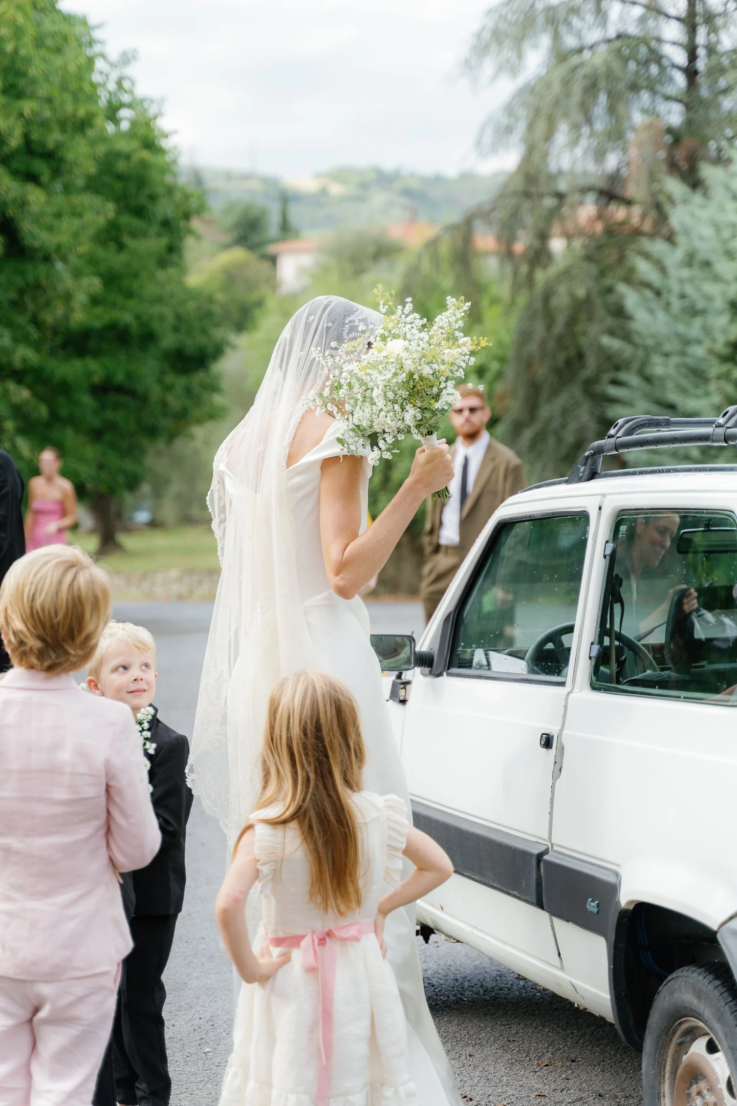 A bride in a white dress and veil holding a bouquet of white flowers, standing next to a white vehicle with children and adults around during a wedding celebration outdoors.