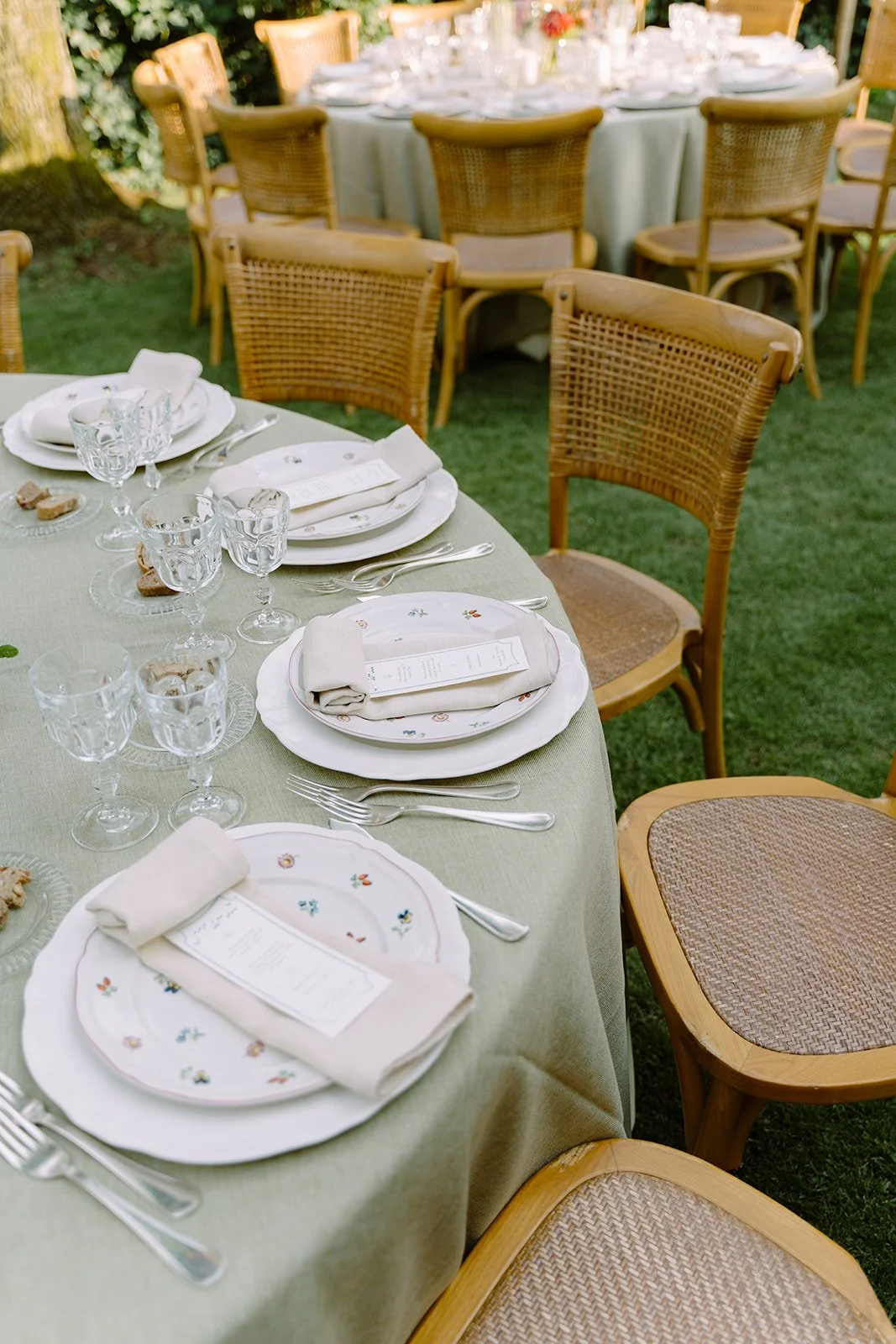 An outdoor dining table set with white plates, wine glasses, silverware, and napkins on a green tablecloth, surrounded by wooden chairs on a grassy area.