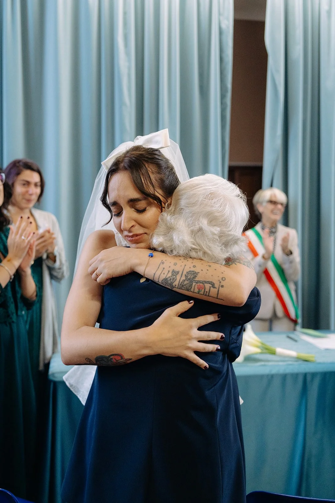 A young woman, wearing a white veil, hugging an older woman with white hair at a formal event, possibly a wedding. Two women in the background are clapping and smiling, with a woman wearing an Italian flag sash.