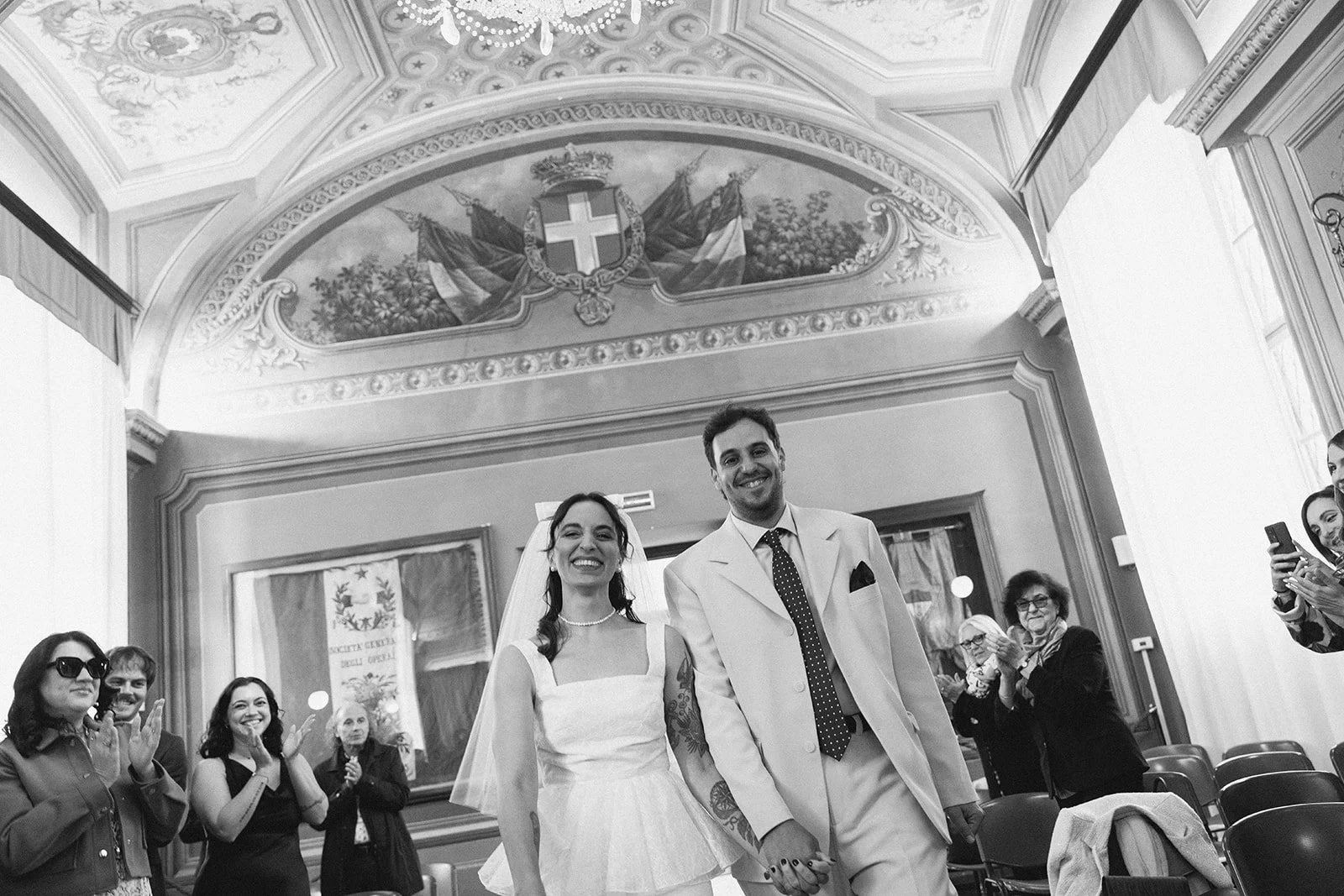 A bride and groom holding hands, smiling, walking down the aisle during their wedding ceremony, with guests clapping and taking photos in a decorated room.