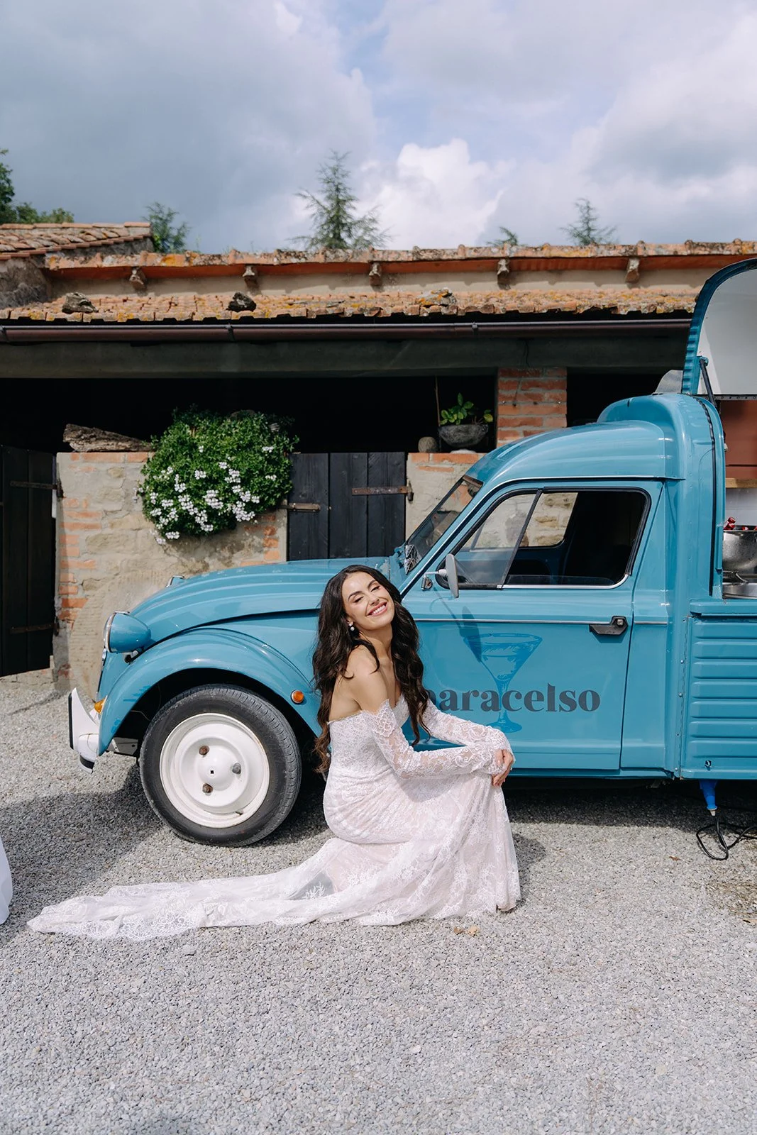 A woman in a white wedding dress sitting in front of a blue vintage van with the text 'aracelso' on the side, outdoors with a rustic building and trees in the background.