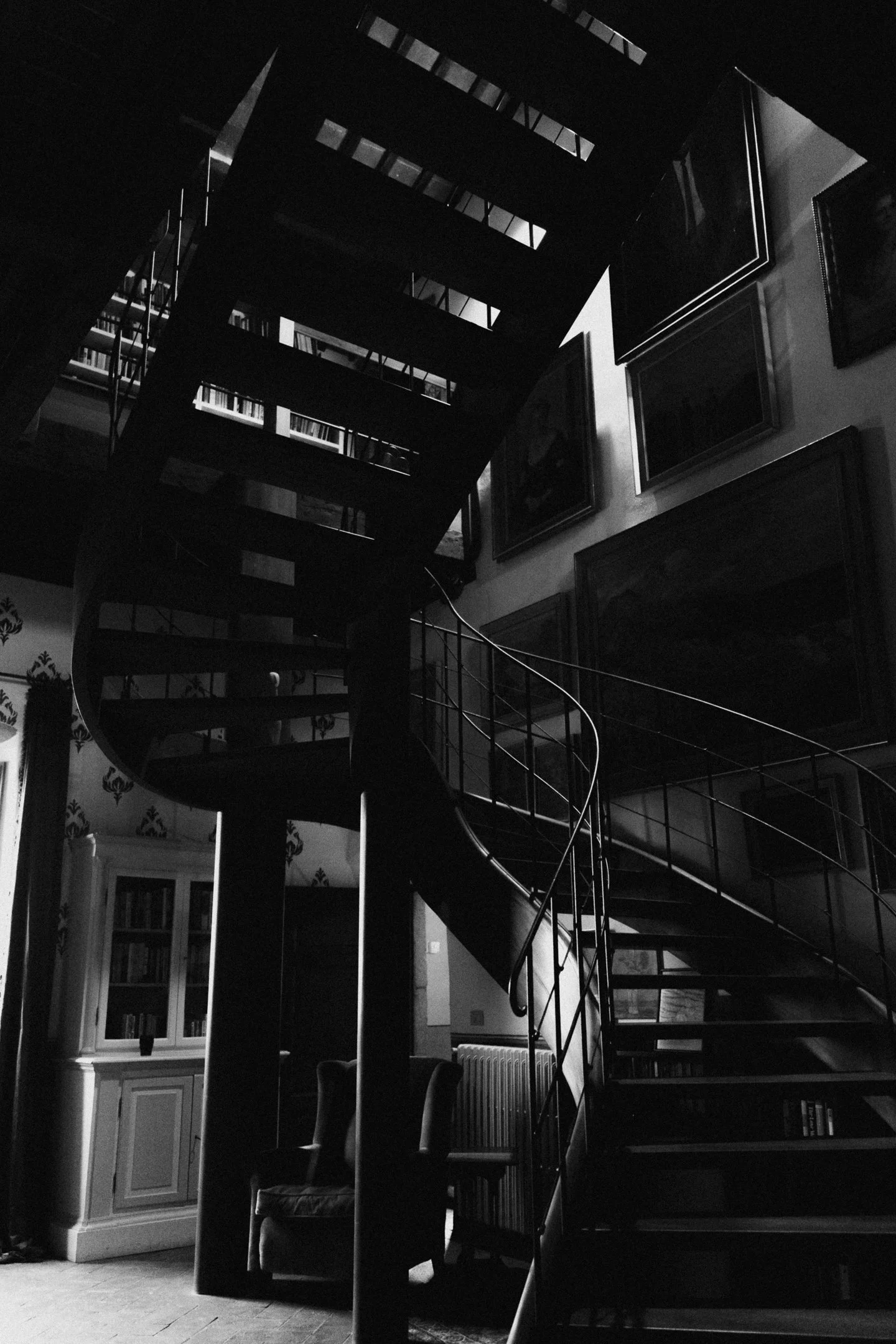 A black and white interior scene featuring a spiral staircase leading to the second floor, with framed artwork on the walls and a bookshelf with glass doors on the left side.