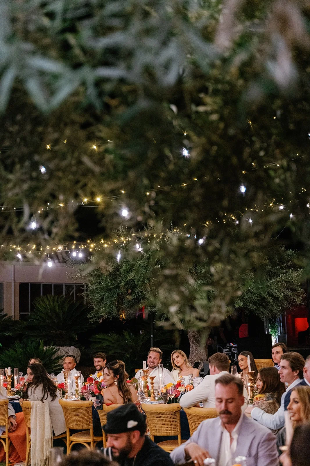 People sitting at banquet tables, smiling and enjoying a celebration, with festive decorations and string lights hanging above in an outdoor setting.