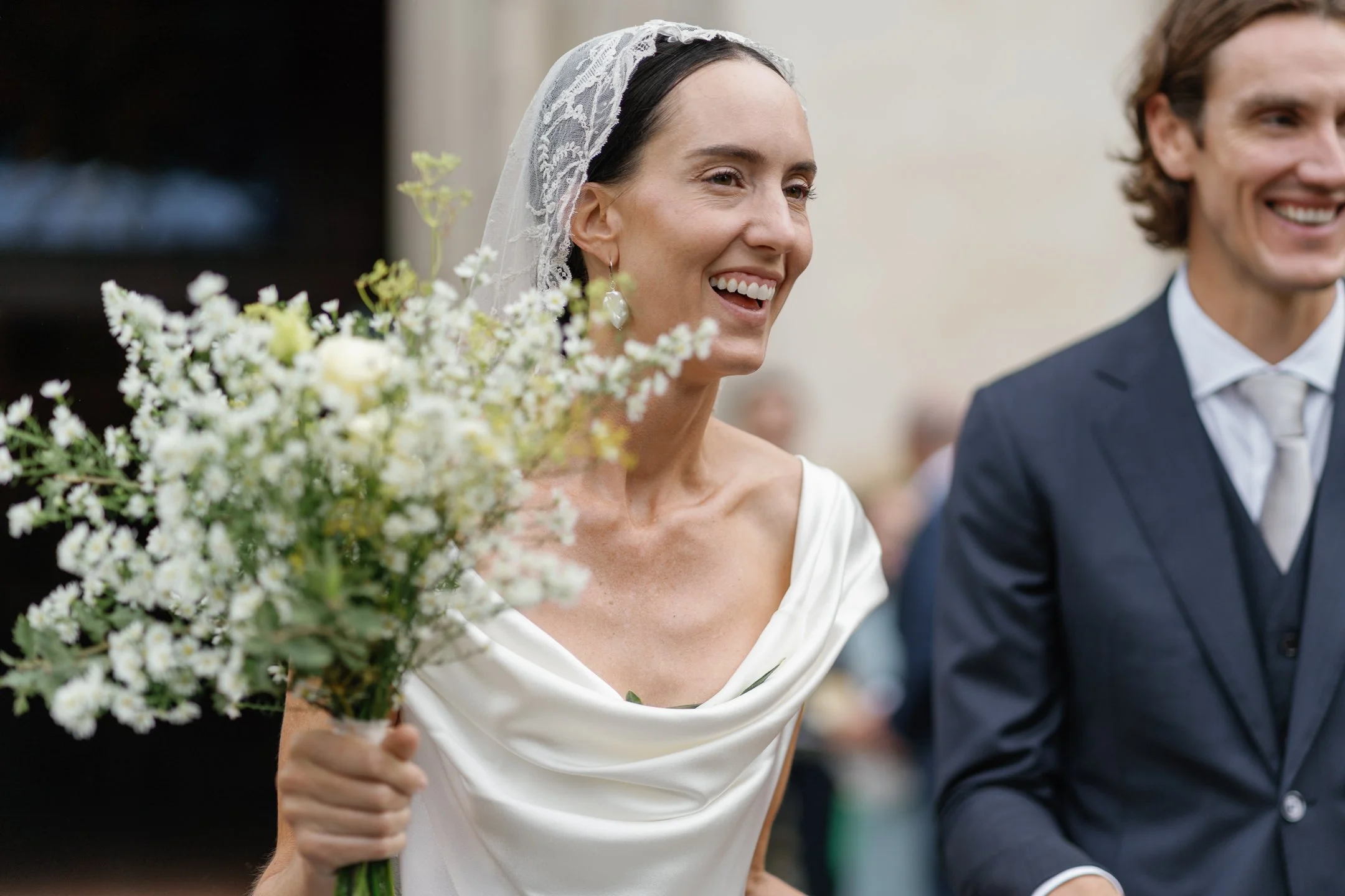 Woman in wedding dress and lace veil holding a bouquet of white flowers, smiling, standing next to a man in a navy suit and light tie at a wedding ceremony.