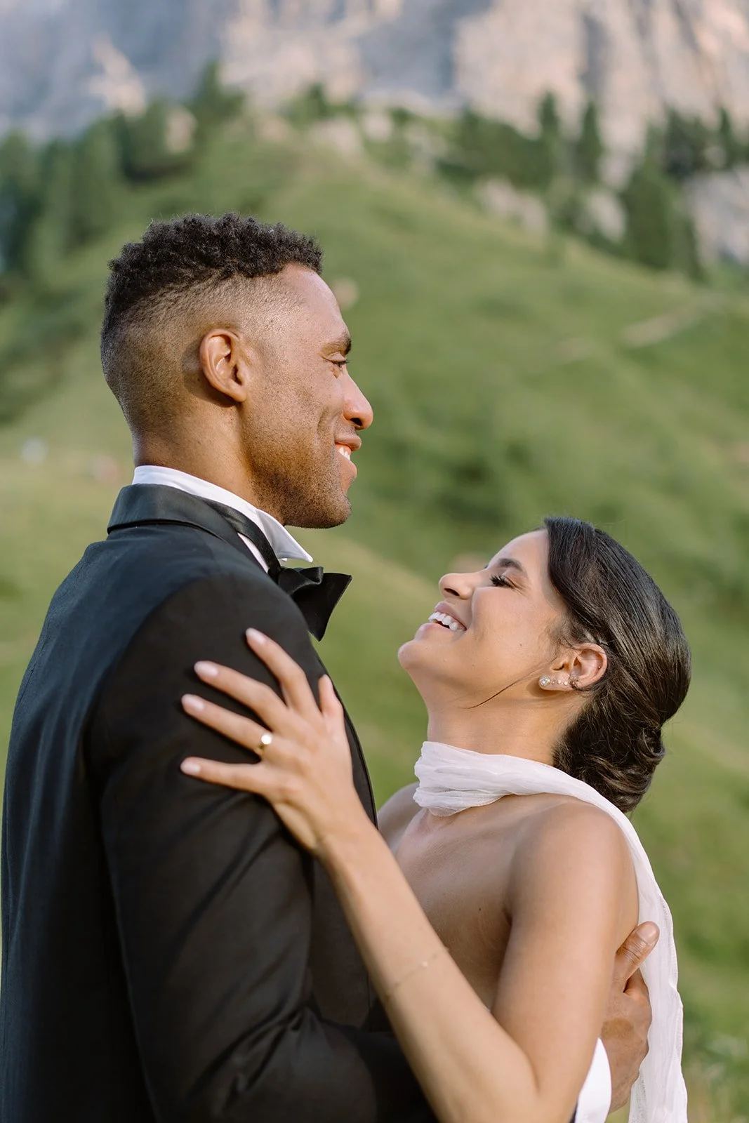 A happy couple in formal attire, smiling and embracing outdoors with a scenic mountain and green landscape in the background.