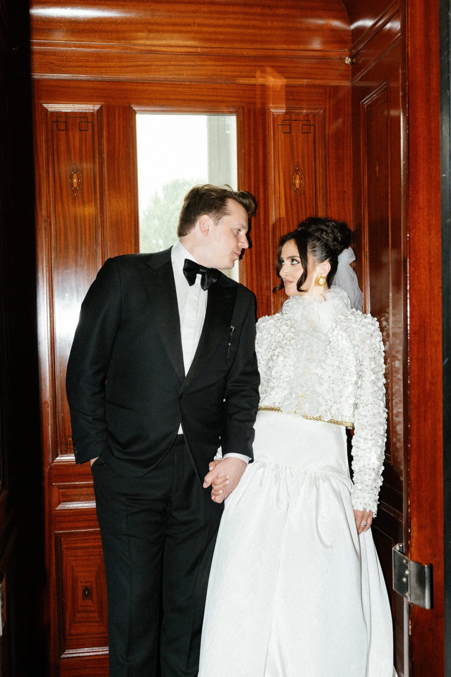 A bride and groom holding hands inside a wooden-paneled elevator with a window behind them.