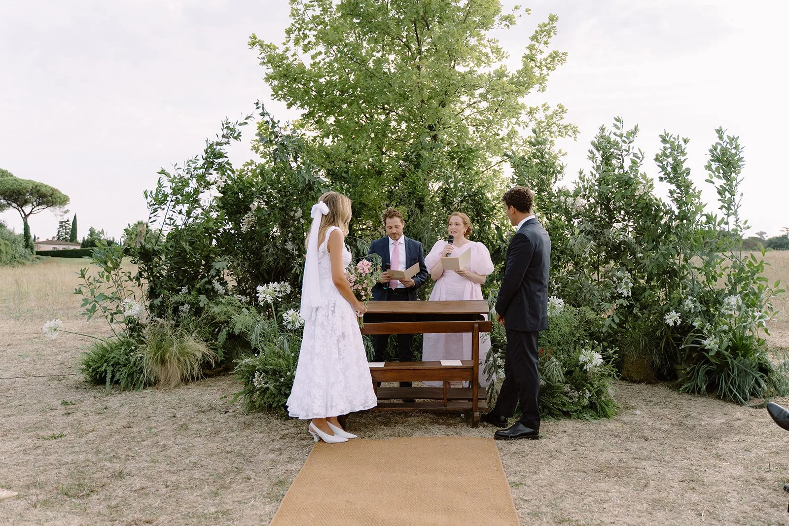 A wedding ceremony taking place outdoors with a bride and groom standing facing each other, with officiants reading from papers. The bride is dressed in a white lace dress holding a bouquet, and the groom is in a dark suit. They are surrounded by gre