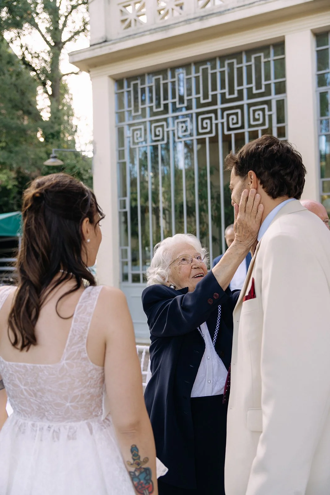 An elderly woman touching the face of a man in a white suit, with a woman with a tattoo on her arm standing nearby, during an outdoor event.