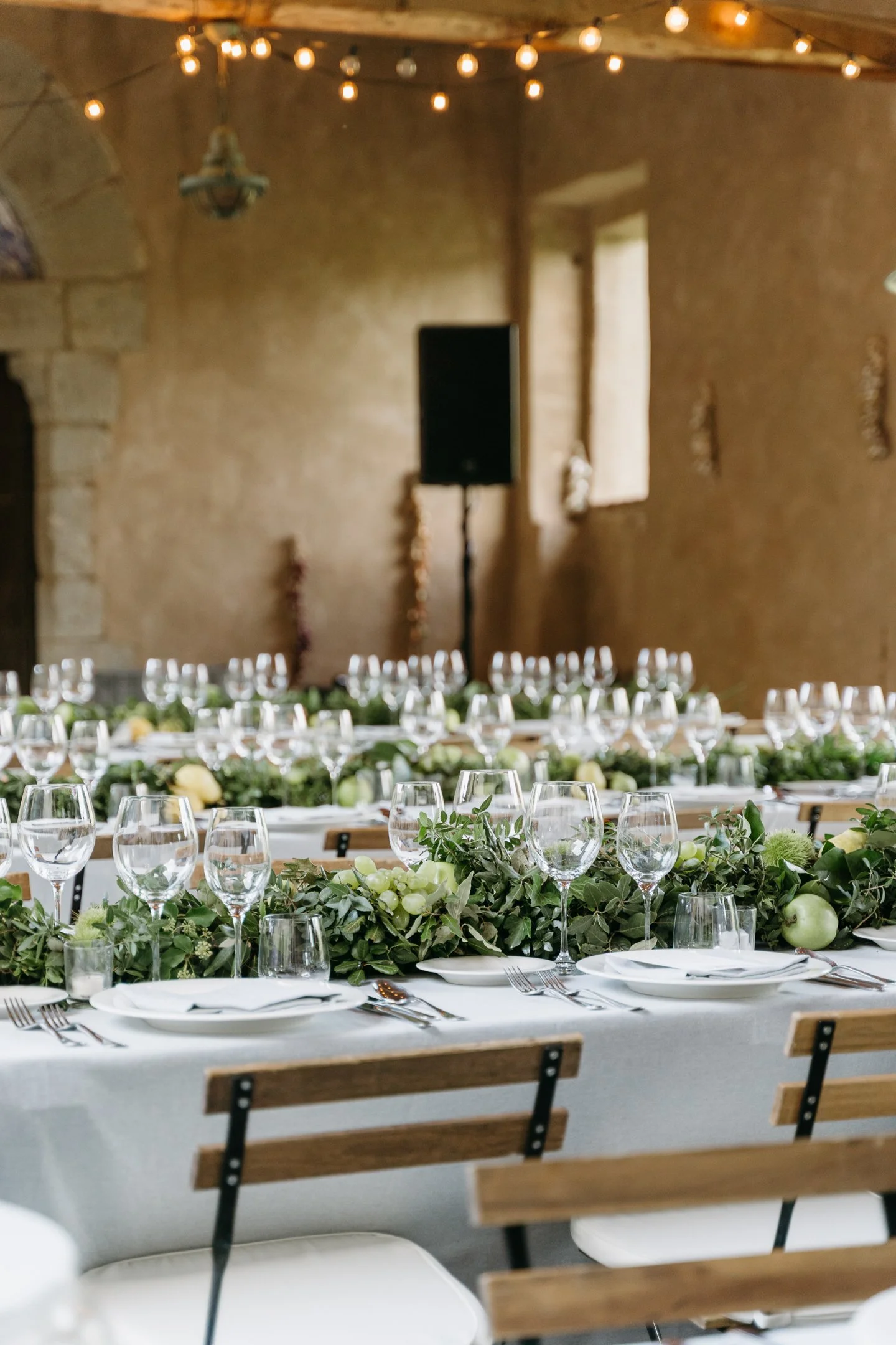 Wedding reception table decorated with greenery, white flowers, and multiple wine glasses, set in a rustic hall with string lights hanging from the ceiling.