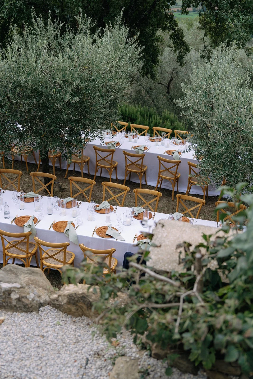 Long outdoor dining table set with white tablecloth, glassware, woven placemats, and green napkins, surrounded by wooden chairs, nestled among trees and rocks in a garden or natural setting.