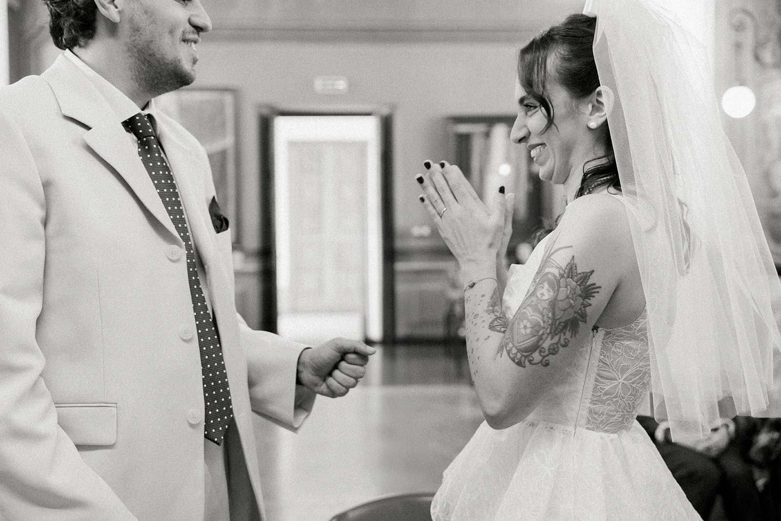A bride and groom smiling at each other during their wedding ceremony, with the bride wearing a veil and a tattoo sleeve on her arm.