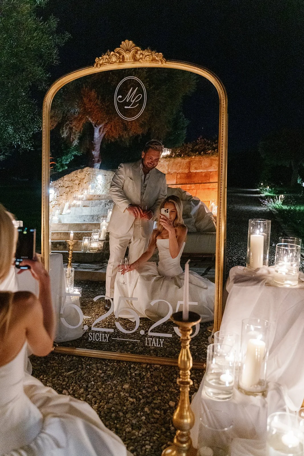 A couple takes a selfie in a mirror during a wedding celebration, with candles and decorations around them at night.