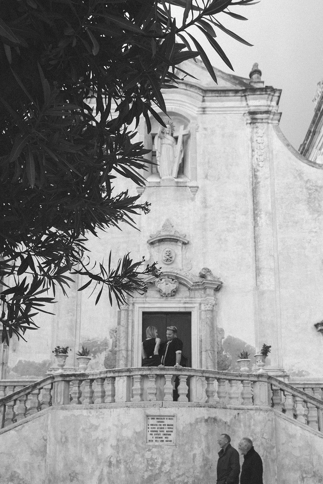 Black and white photo of a church facade with a balcony. Two people are standing on the balcony talking. Two men are walking below the balcony. A large tree with leaves partially covers the upper left corner of the photo.