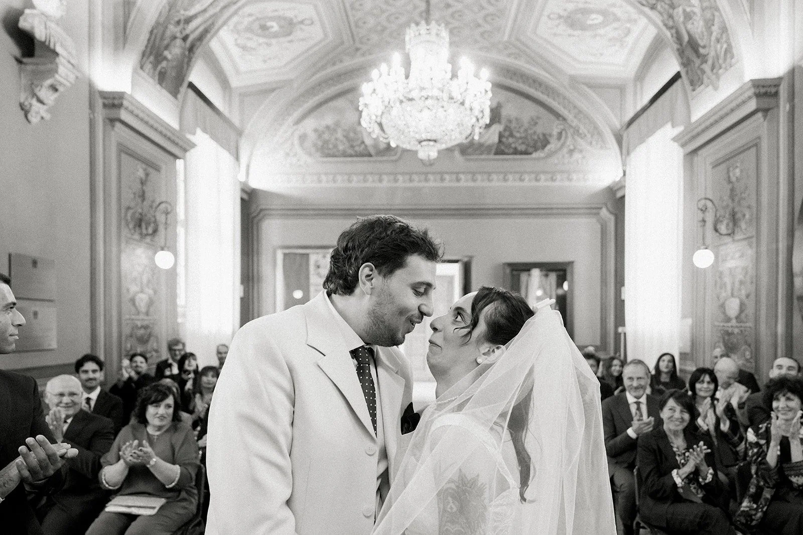 Black and white photo of a wedding ceremony in a grand, ornate hall, with a bride and groom gazing into each other's eyes, surrounded by seated guests applauding.