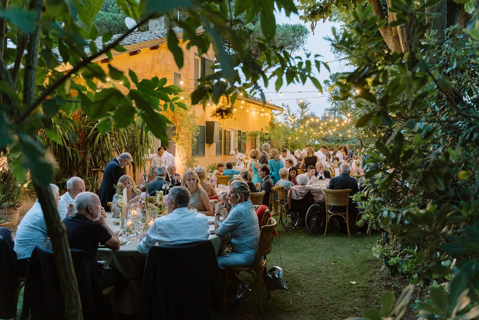 Outdoor evening dinner gathering with guests seated at tables under string lights in a garden with greenery and a rustic building in the background.
