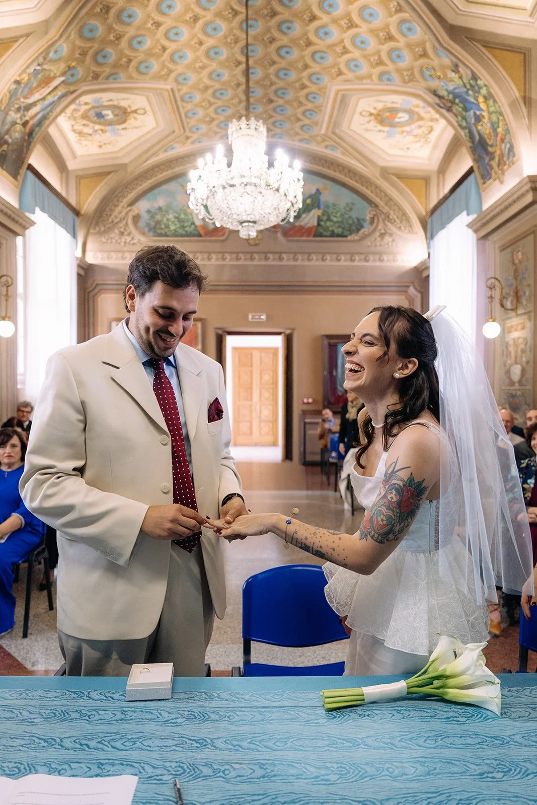 A bride and groom exchanging rings during a wedding ceremony inside a decorated hall with a high, ornate ceiling and a chandelier. The bride has tattoos and is smiling, and the groom is in a light-colored suit with a red polka dot tie.