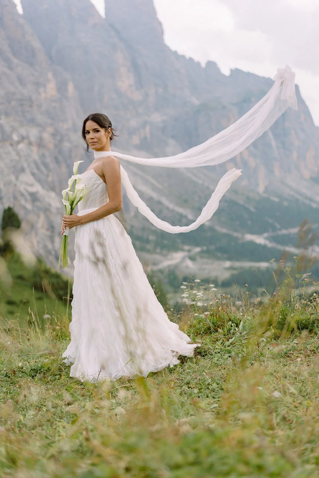 A woman in a white wedding dress holding a bouquet of calla lilies outdoors with mountains and grassy terrain in the background.