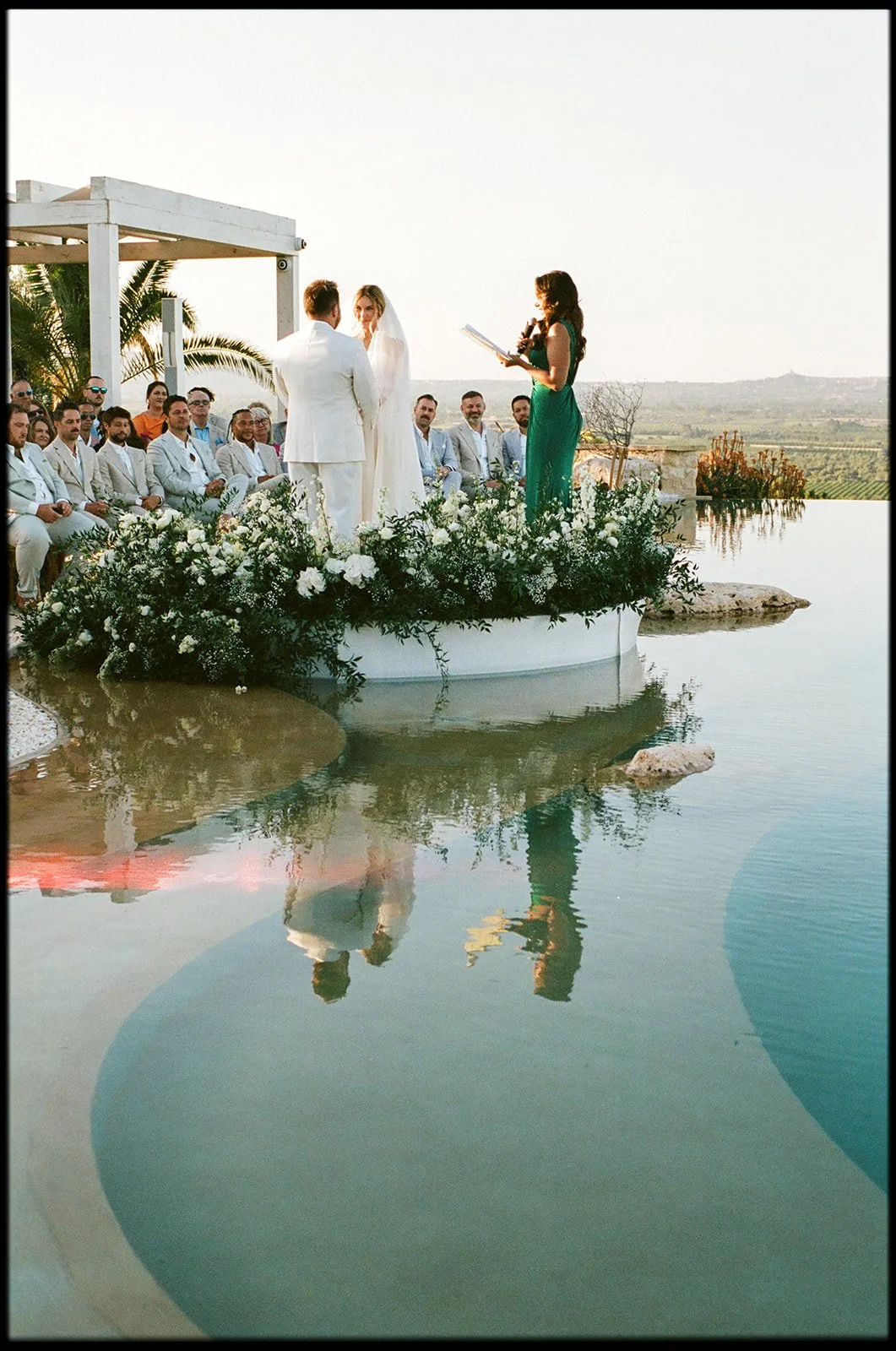 A wedding ceremony taking place outdoors on a platform above water, with guests seated and officiant reading from a book.