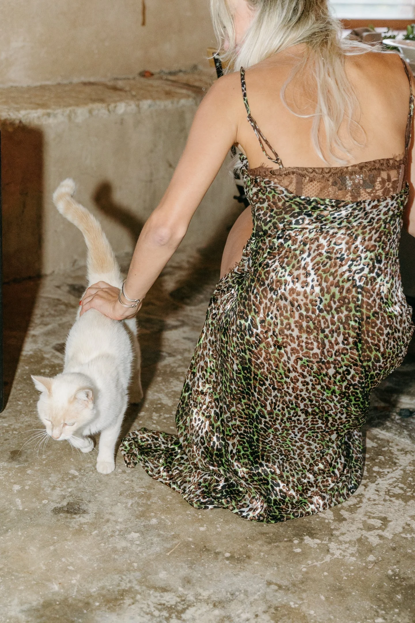 A woman with blonde hair wearing a leopard print dress is kneeling on a concrete floor, petting a white cat with a curled tail.