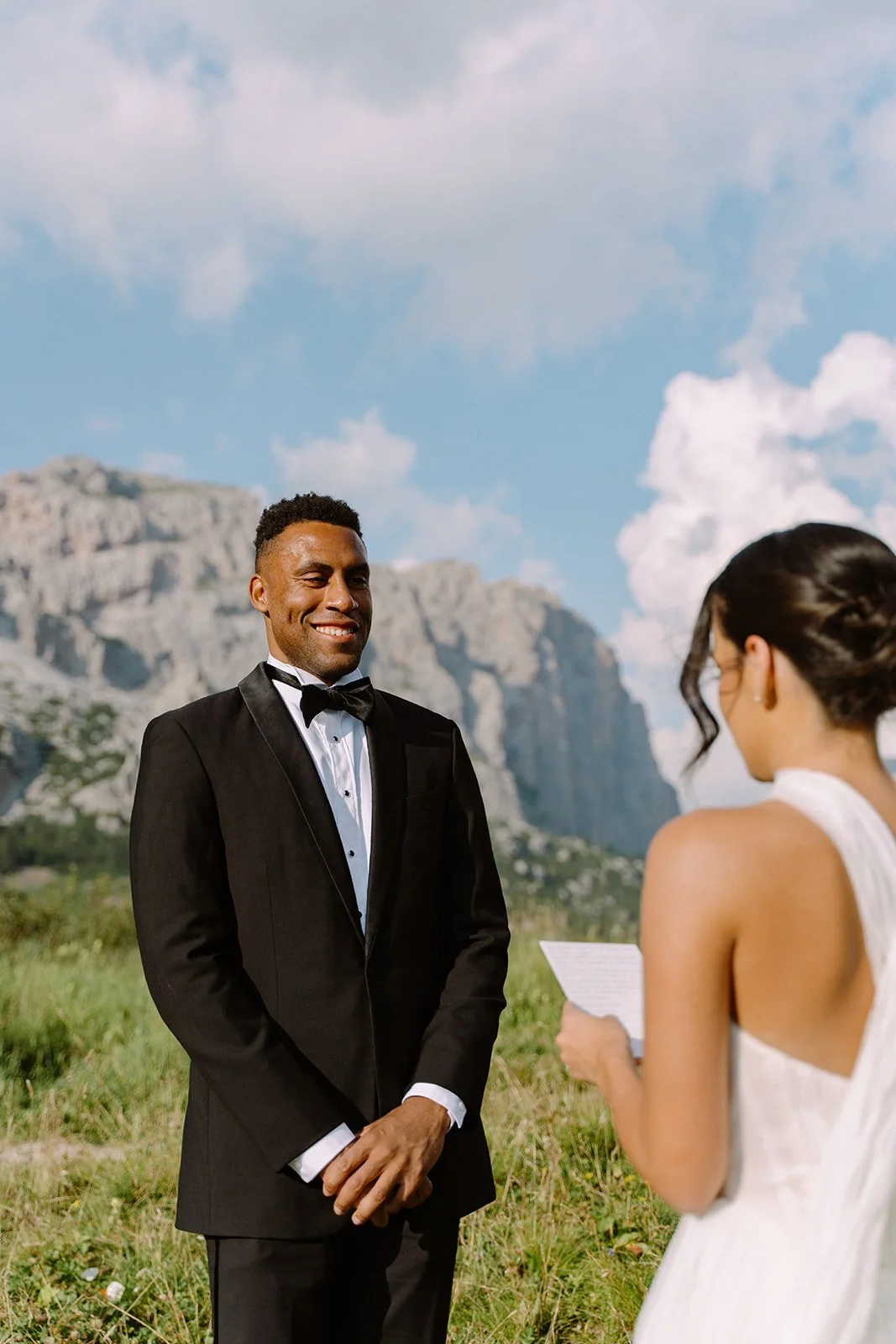 A couple is exchanging wedding vows outdoors, with a mountain and blue sky background. The groom is wearing a tuxedo, and the bride is in a white dress holding a paper.