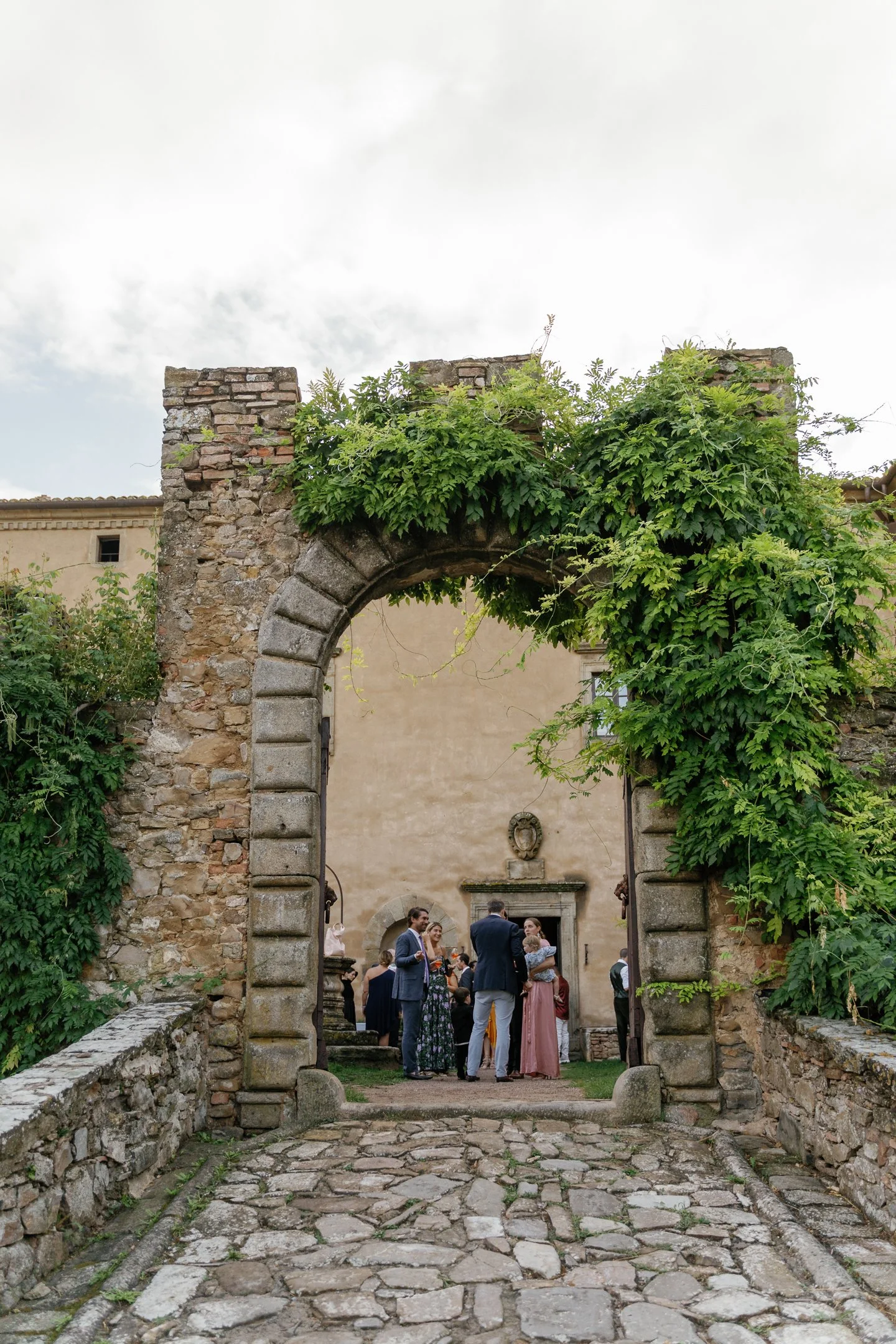 A group of people gathered under an ancient stone archway covered in green vines, with a beige building in the background, possibly during a wedding or social event.