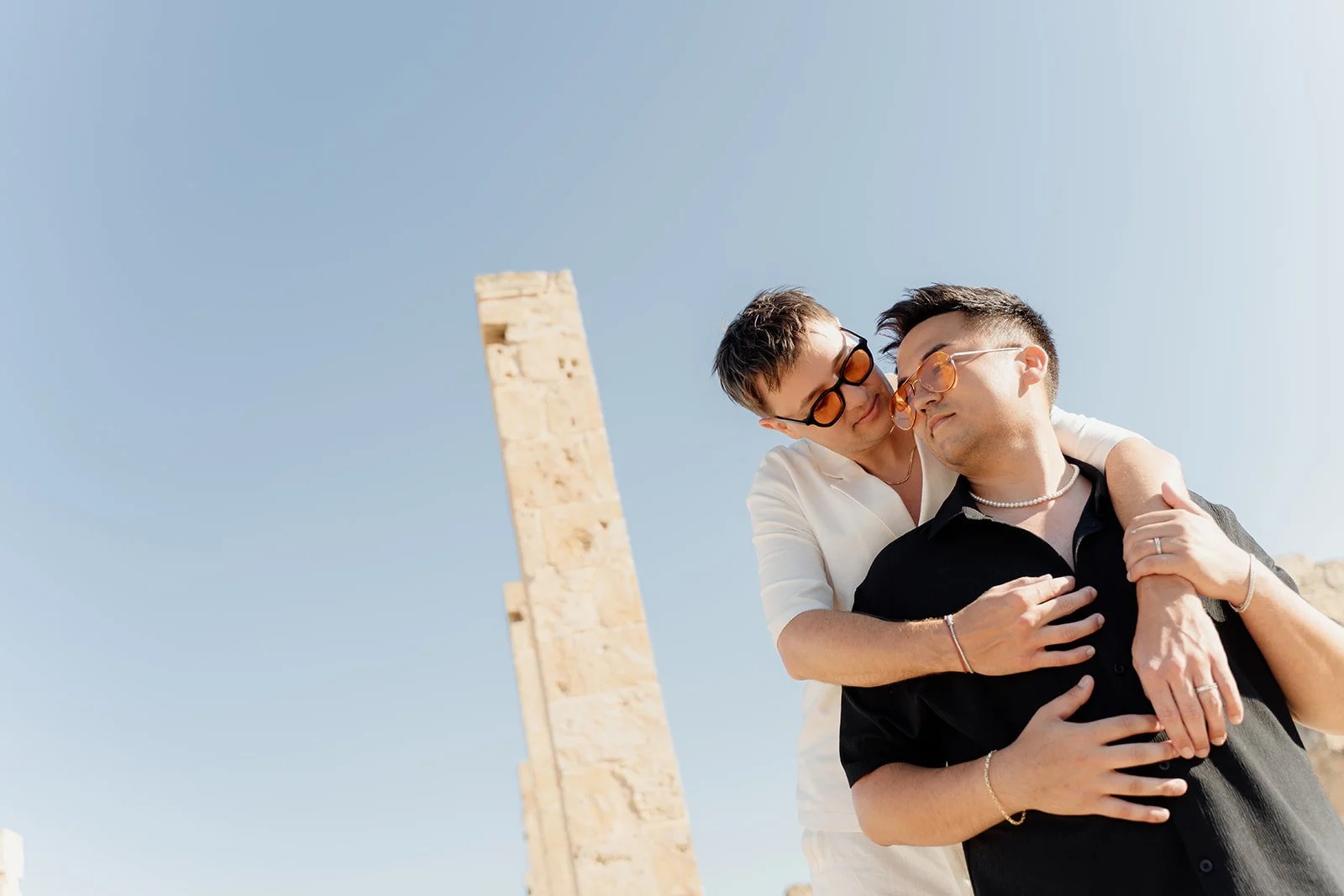A couple wearing glasses under a clear blue sky, with ancient ruins in the background, embracing each other lovingly.