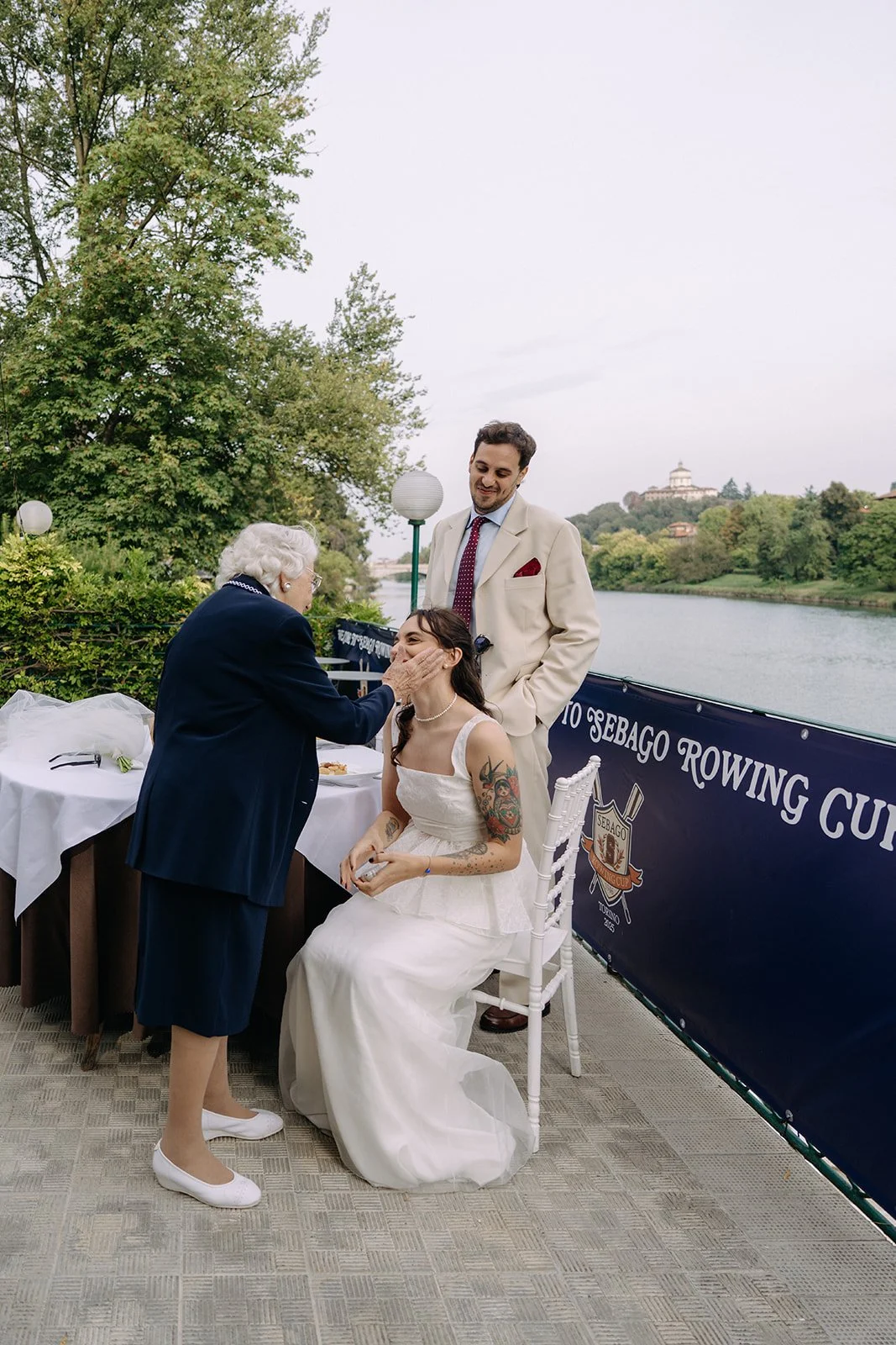 A woman in a wedding dress with tattoos on her arms sits on a white chair, smiling as an elderly woman touching her face, while a man in a cream suit standing behind her looks on, on a riverfront terrace with trees and a building in the background du