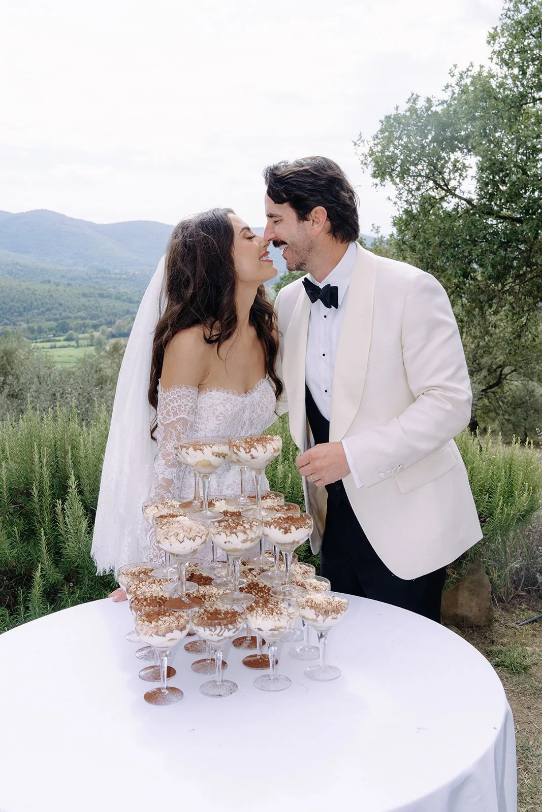 A newlywed couple smiling and leaning close to each other outdoors, with a mountain landscape and greenery in the background, standing behind a table with a pyramid of dessert cups topped with whipped cream and cocoa powder.