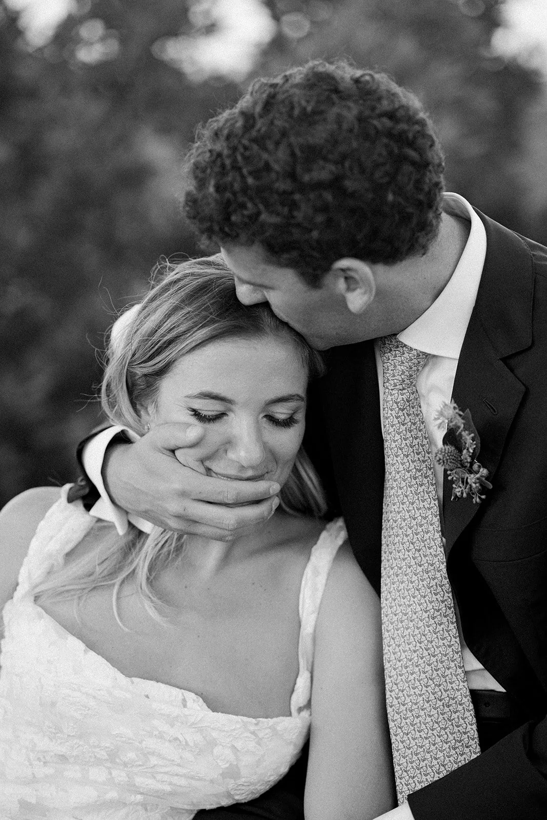 A black and white photograph of a man gently kissing a woman on her forehead while holding her chin, both with closed eyes, in an outdoor setting.