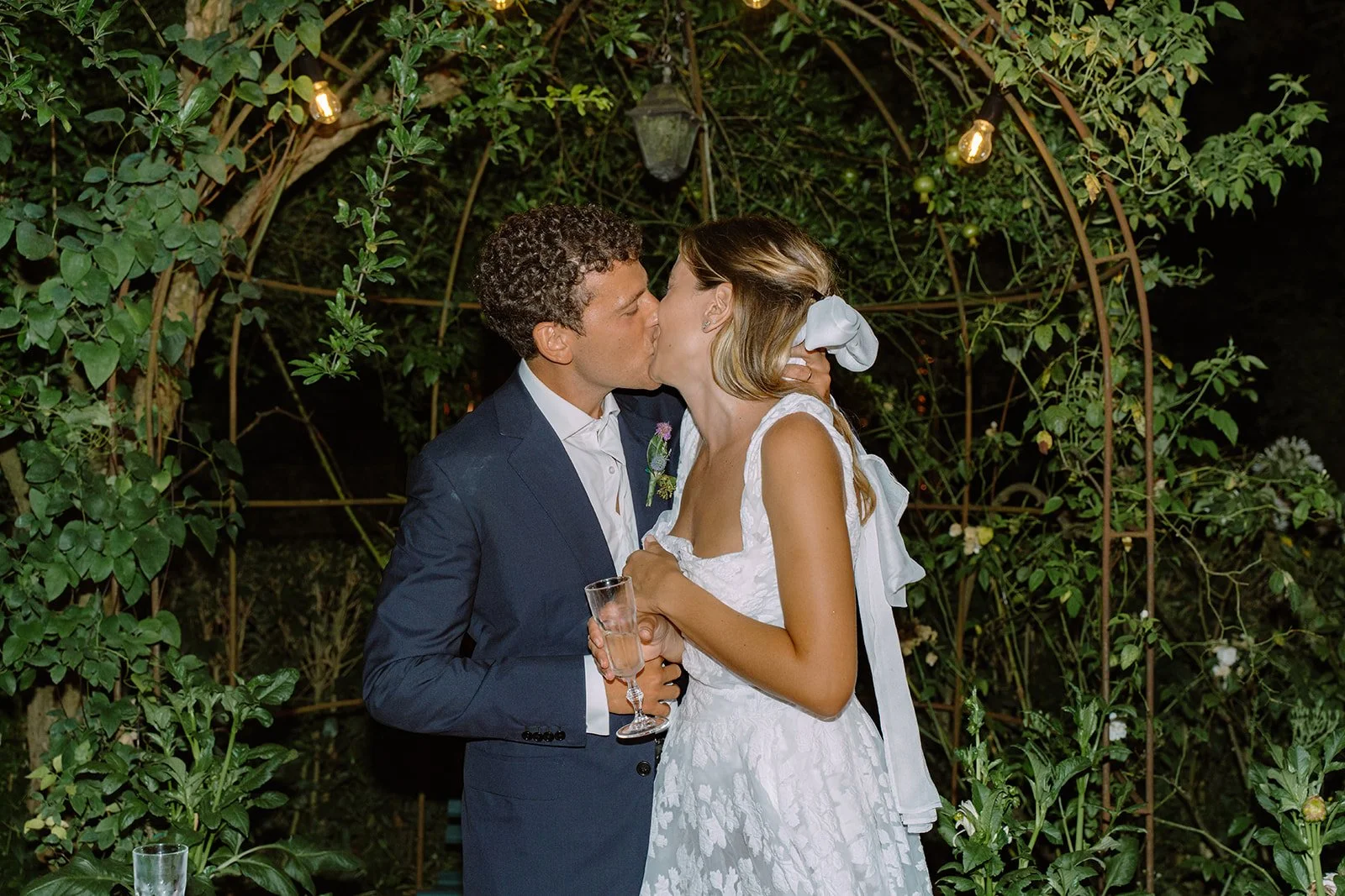 Newlywed couple sharing a kiss at their wedding reception outdoors, with greenery and string lights in the background.